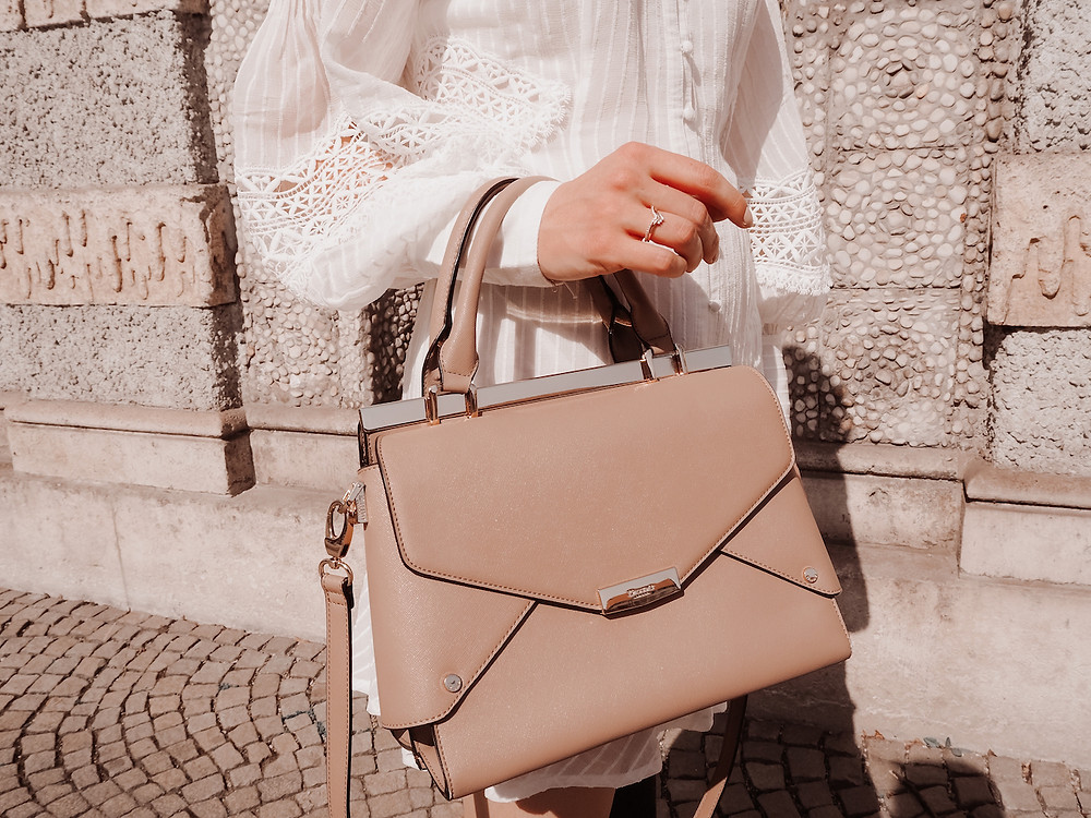 Young woman wearing an outfit consisting of a white lace dress, white coat, brown boots, and a brown bag, standing in front of a stone wall