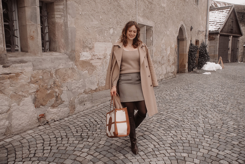 Junge Frau in einem beigen Mantel, mit großer Canvas Tasche und braunen Stiefeln vor einer Sandsteinwand