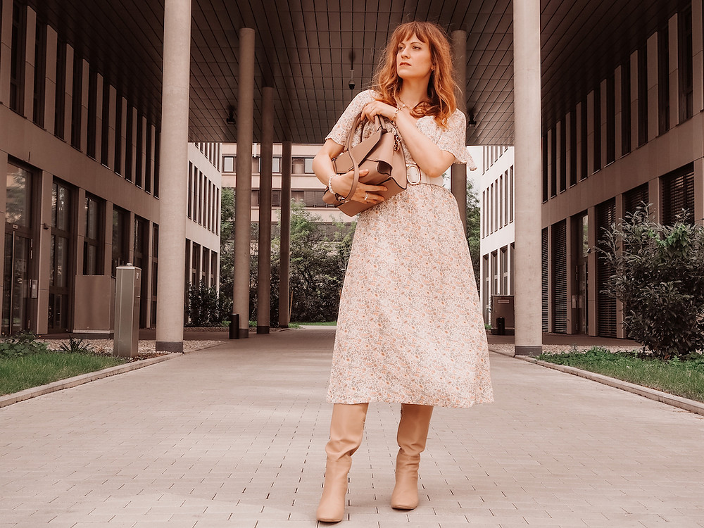 Young woman dressed in a floral midi dress, broad waist belt, beige boots and a brown bag in front of an office building
