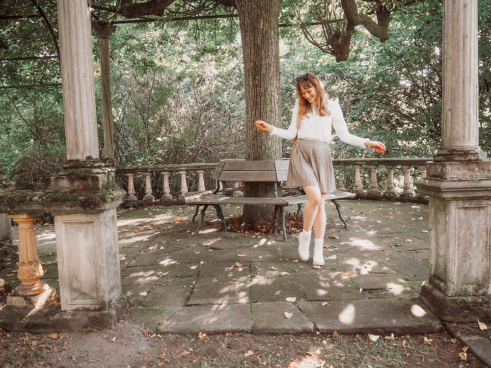 Young woman wearing a pleated mini skirt, trench coat, white lace blouse, white lace-up boots and Strathberry bag in a park