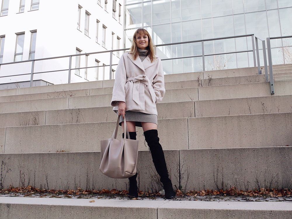 Girl dressed in beige wool jacket, grey jumper dress, black over the knee boots and beige shopper bag standing on stairs
