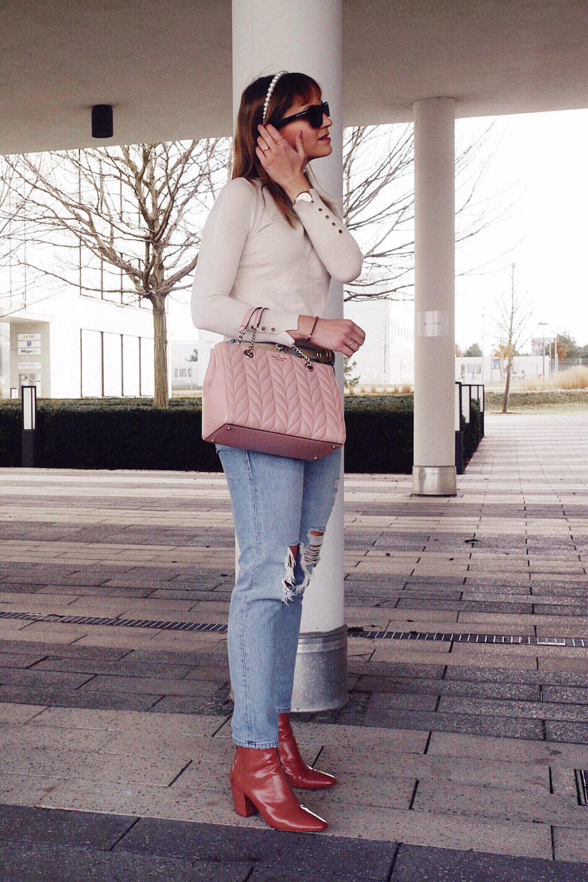 Girl in cream roll neck and distressed denim with pink boots and Kate Spade bag in front of a window