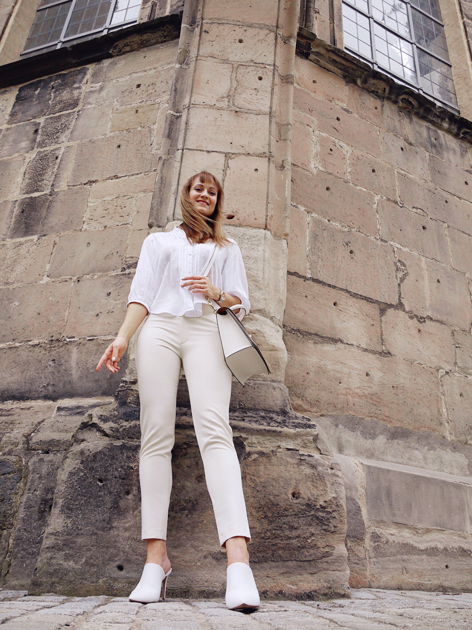 Junge Frau in einem Outfit aus weißer Bluse, cremefarbener Hose und weißen, spitzen Schuhen unter einem Steinbogen