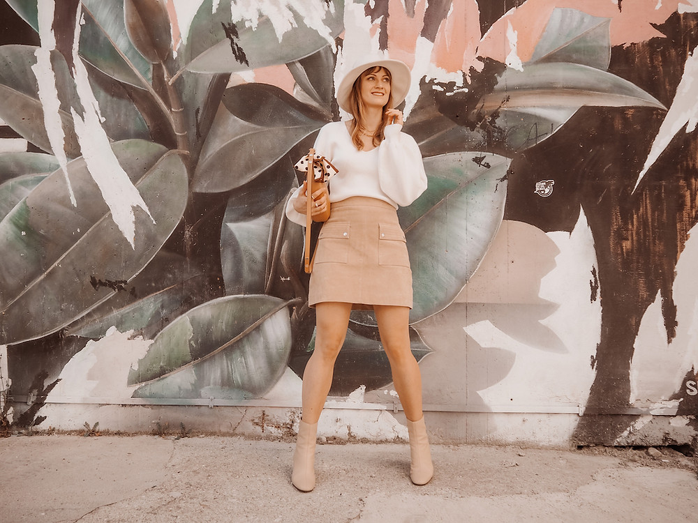 Junge Frau in einem Outfit aus Trenchcoat, Wildleder-Rock und weißem Pullover vor einer Graffiti-Wand
