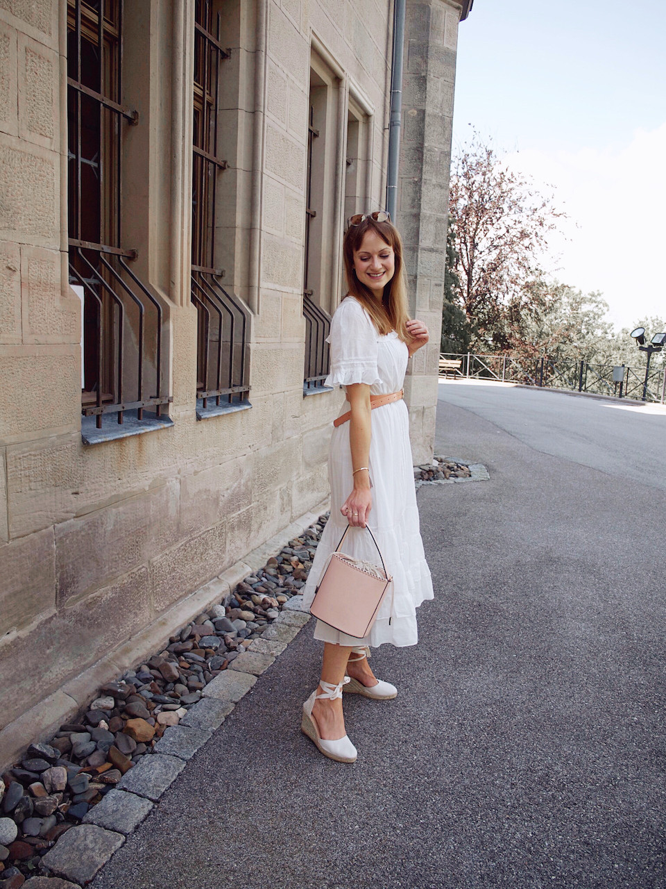 Young woman dressed in a midi length white summer dress, waist belt, white wedges, and pink bucket bag in front of a stone building