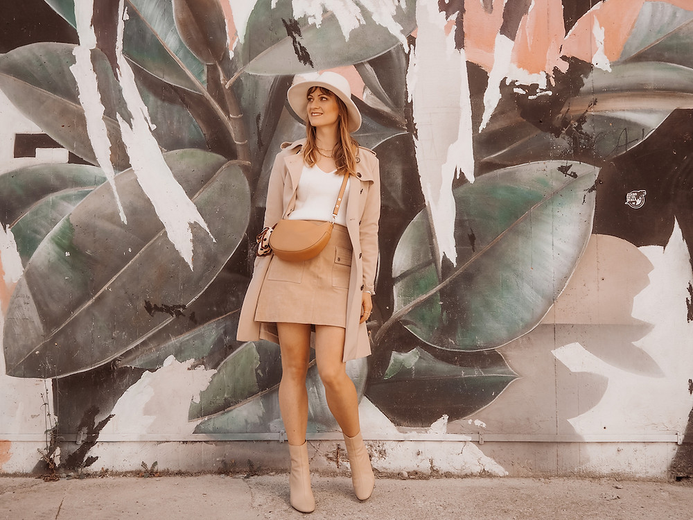 Junge Frau in einem Outfit aus Trenchcoat, Wildleder-Rock und weißem Pullover vor einer Graffiti-Wand