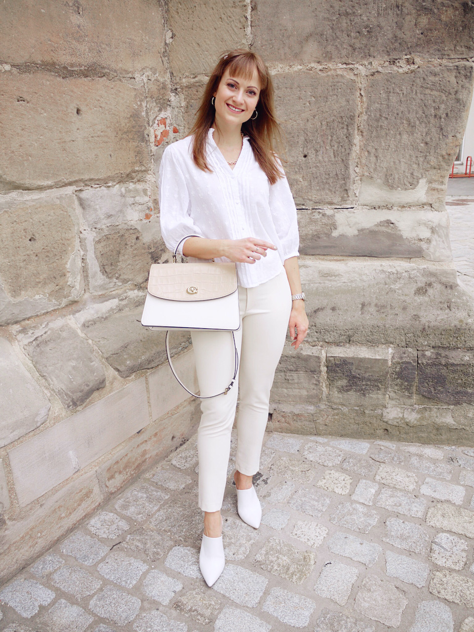 Young women dressed in white blouse, cream colored trousers, and white mules carrying a Coach bag, standing in front of a stone wall
