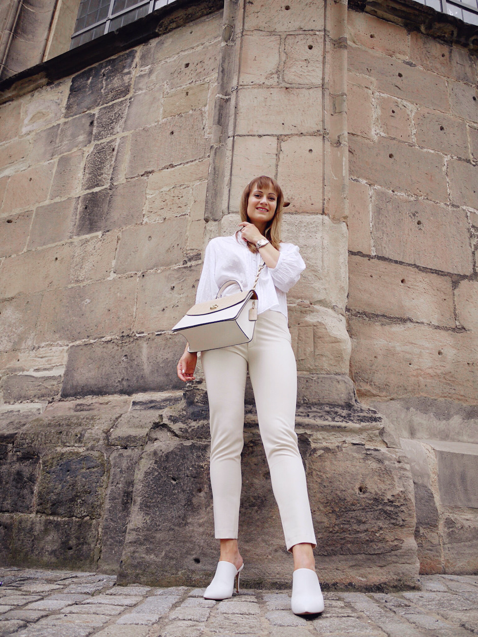 Young women dressed in white blouse, cream colored trousers, and white mules carrying a Coach bag, standing under a stone archway