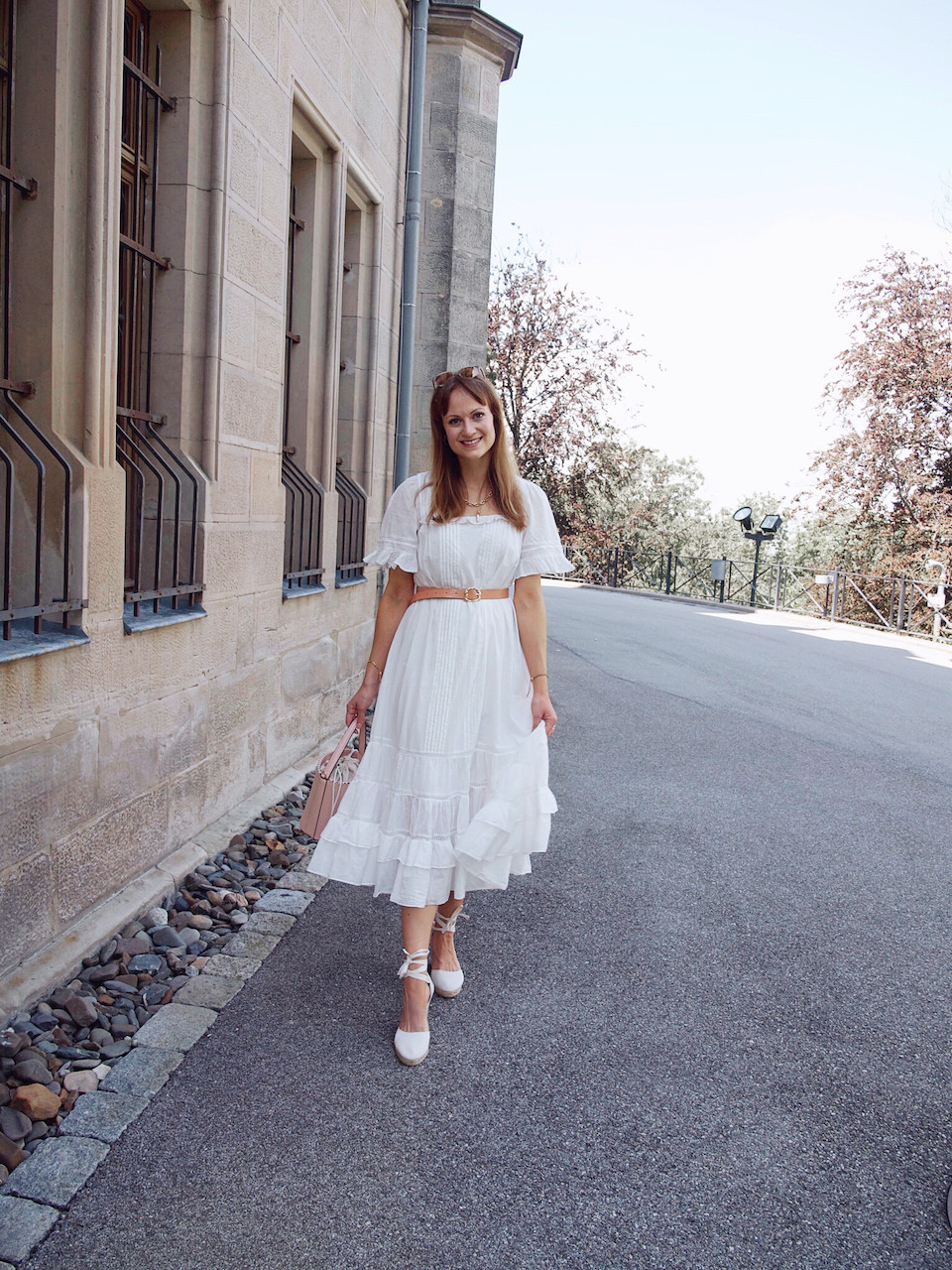 Young woman dressed in a midi length white summer dress, waist belt, white wedges, and pink bucket bag in front of a stone building