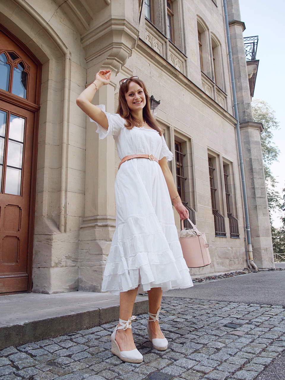 Junge Frau in einem weißen Sommerkleid in Midilänge, weißen Keilabsatzschuhen und rosa Beuteltasche vor einem Steingebäude