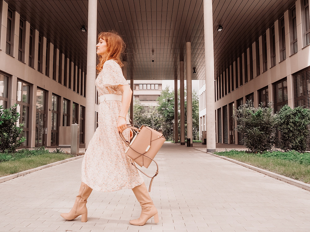 Young woman dressed in a floral midi dress, broad waist belt, beige boots and a brown bag in front of an office building