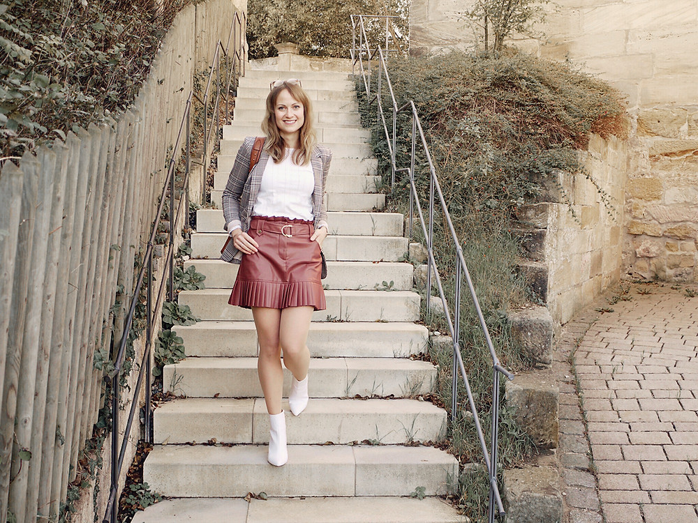 Girl in 70s inspired outfit wearing a faux leather mini skirt, white shirt, checked blazer and white boots walking down steps