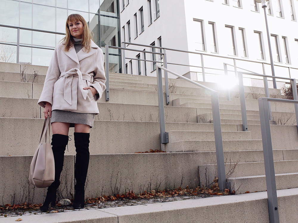 Girl dressed in beige wool jacket, grey jumper dress, black over the knee boots and beige shopper bag standing on stairs