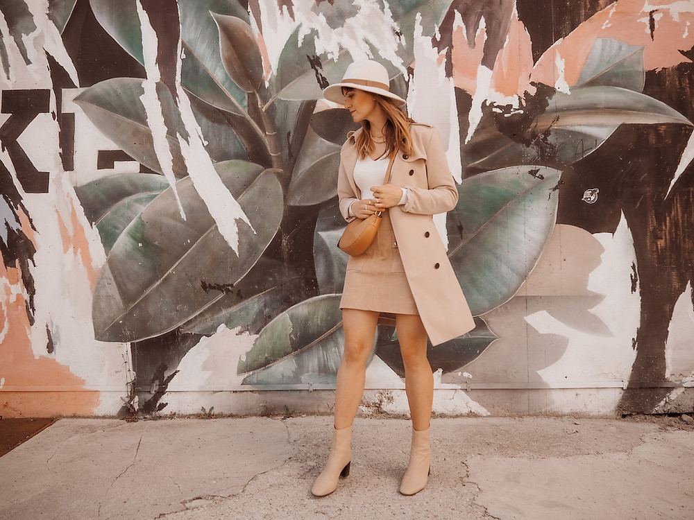 Young woman dressed in a trench coat, suede skirt, and white knit in front of a graffiti wall