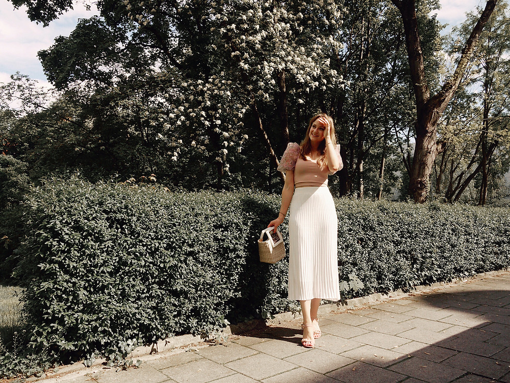 Girl wearing a cream midi skirt, puff sleeve floral top, straw bag, and heeled sandals