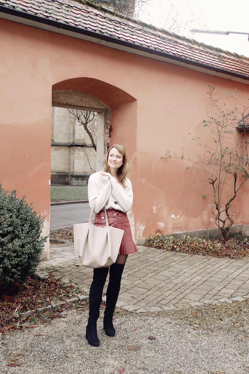 Girl in cozy cardigan, leather mini skirt, over the knee boots and with a big shopper bag standing in front of a gate