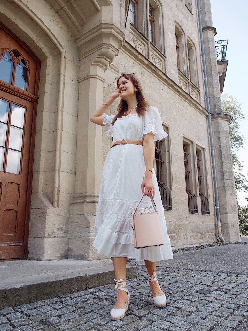 Junge Frau in einem weißen Sommerkleid in Midilänge, weißen Keilabsatzschuhen und rosa Beuteltasche vor einem Steingebäude