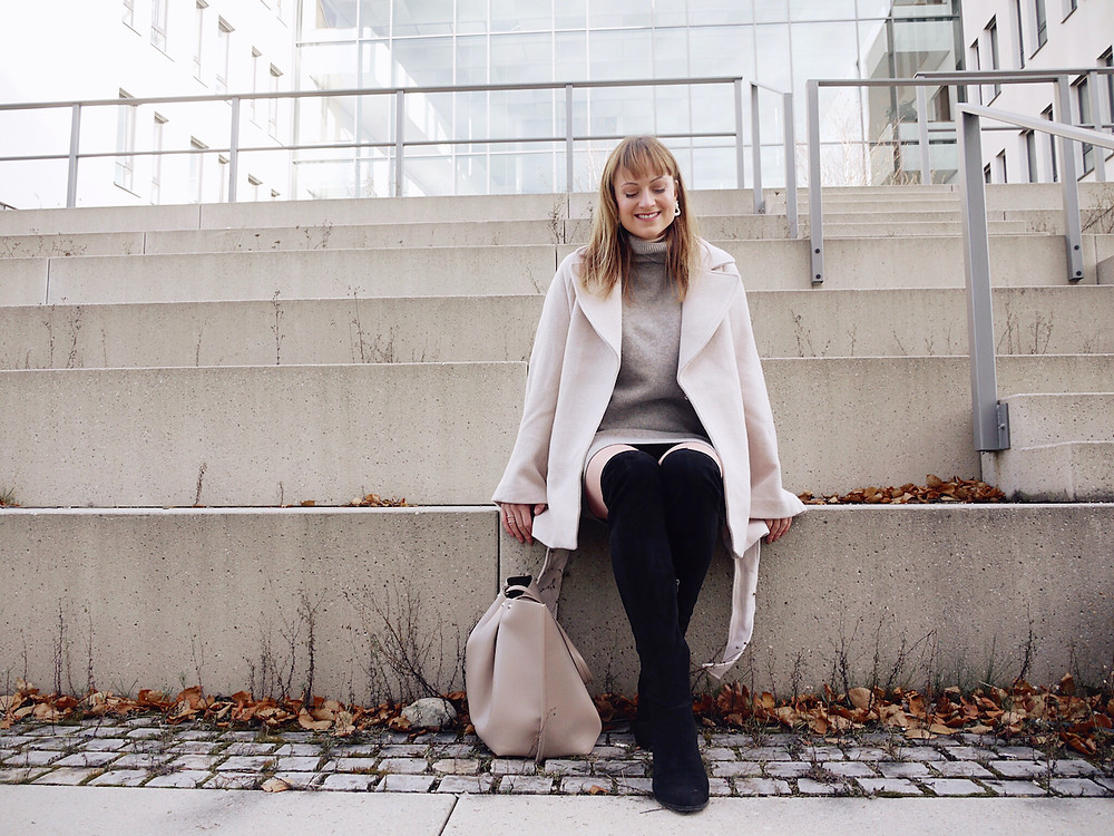 Girl dressed in beige wool jacket, grey jumper dress, black over the knee boots and beige shopper bag standing on stairs