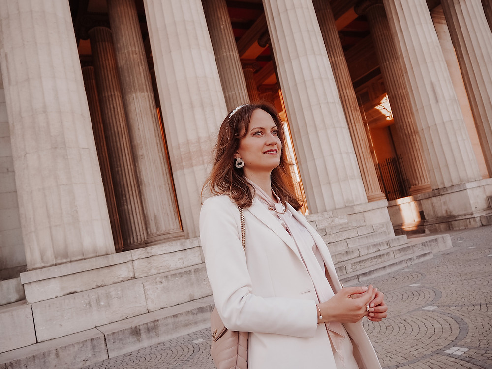 Young woman dressed in an outfit of a blazer, shorts, a silk blouse, and strappy sandals, standing in front of columns