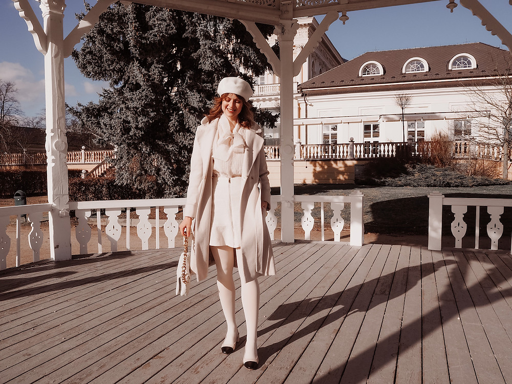 Young woman in an all-cream outfit consisting of a coat, a bouclé mini skirt, a pussy bow blouse, tights, a beret and slingback pumps in a pavillon