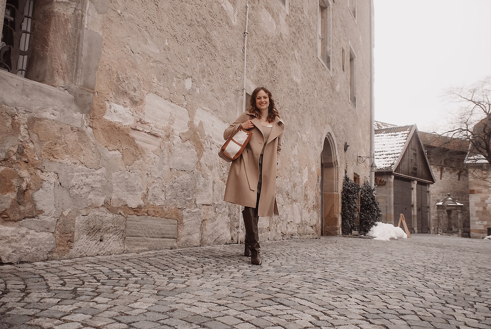 Young woman dressed in a camel trench coat, brown boots and carrying a canvas tote in front of a stone wall