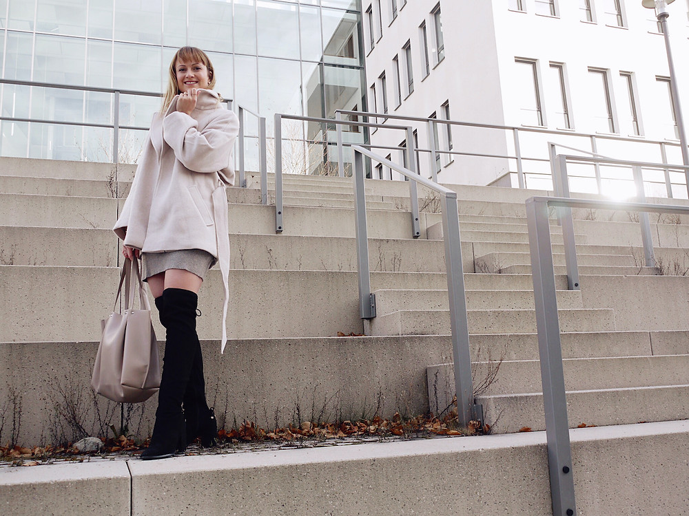 Junge Frau in beige Wolljacke, grauem Pulloverkleid, schwarzen Overknee-Stiefeln und beige Shopper-Tasche auf Stufen