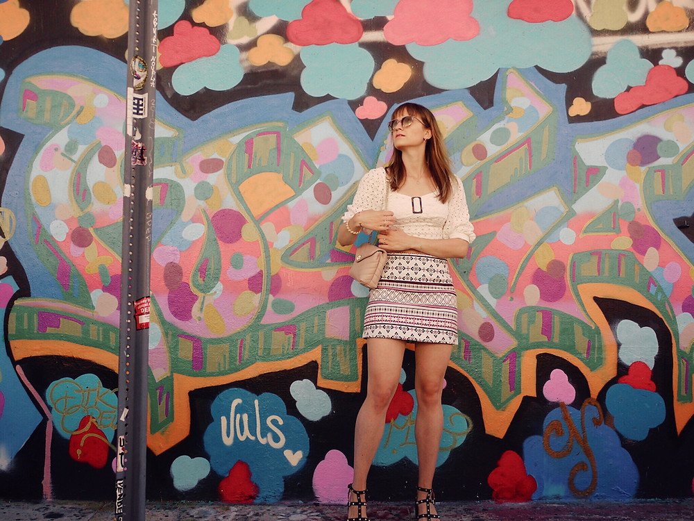 Girl in white lace crop top, patterned skirt, and studded sandals standing in front of a graffitti in Wynwood, Miami