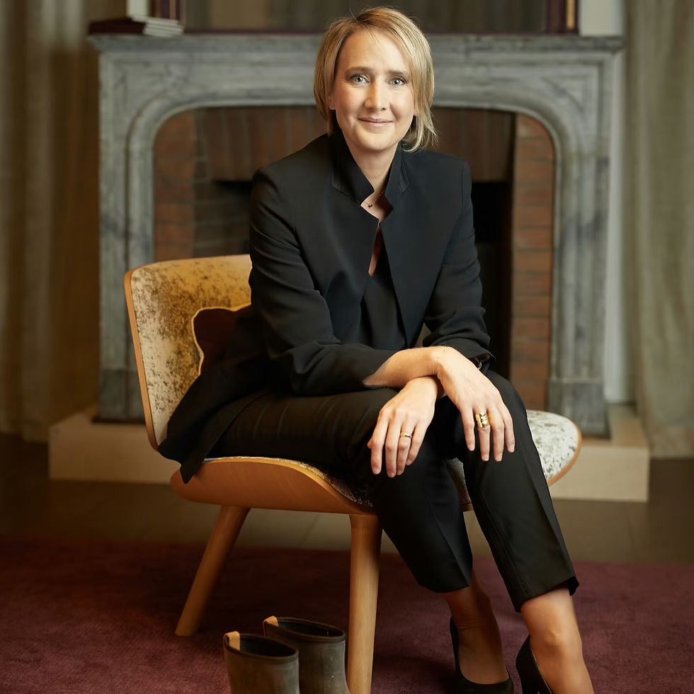 Smiling woman in a black suit sits on a chair in a cozy room with a fireplace. Brown shoes on a maroon carpet enhance the warm setting.