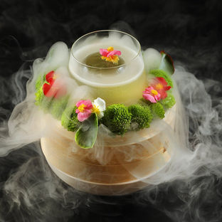 A drink in a glass sits on a wooden base, surrounded by colorful flowers and green moss, with mist enveloping it against a dark background.
