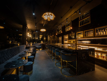 Luxurious bar interior with dark velvet seating, gold accents, and illuminated liquor shelves. Warm ambient lighting creates an elegant mood.