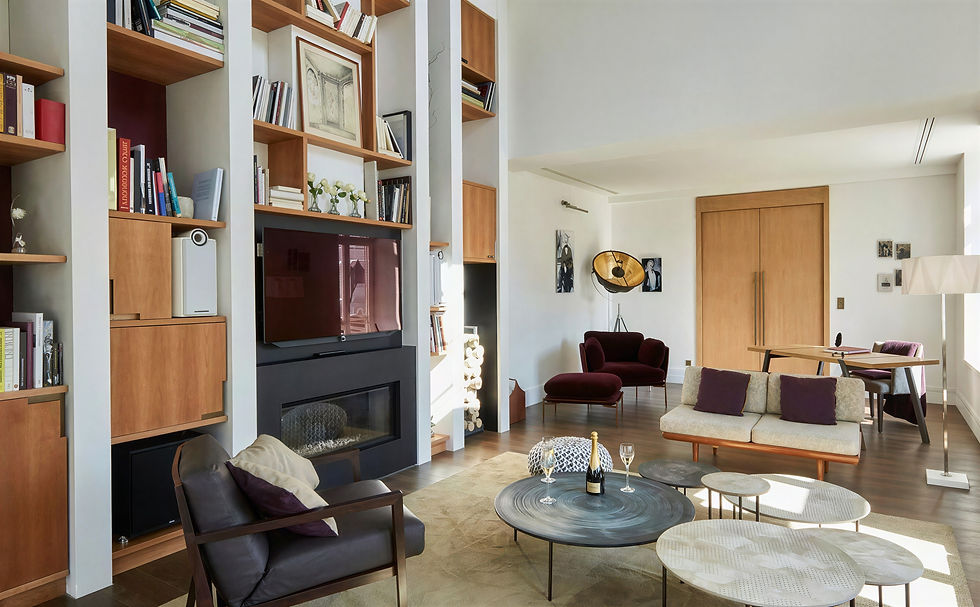 Modern living room with wooden shelves, a red armchair, round tables holding wine, and large artwork. Bright, cozy, and inviting space.