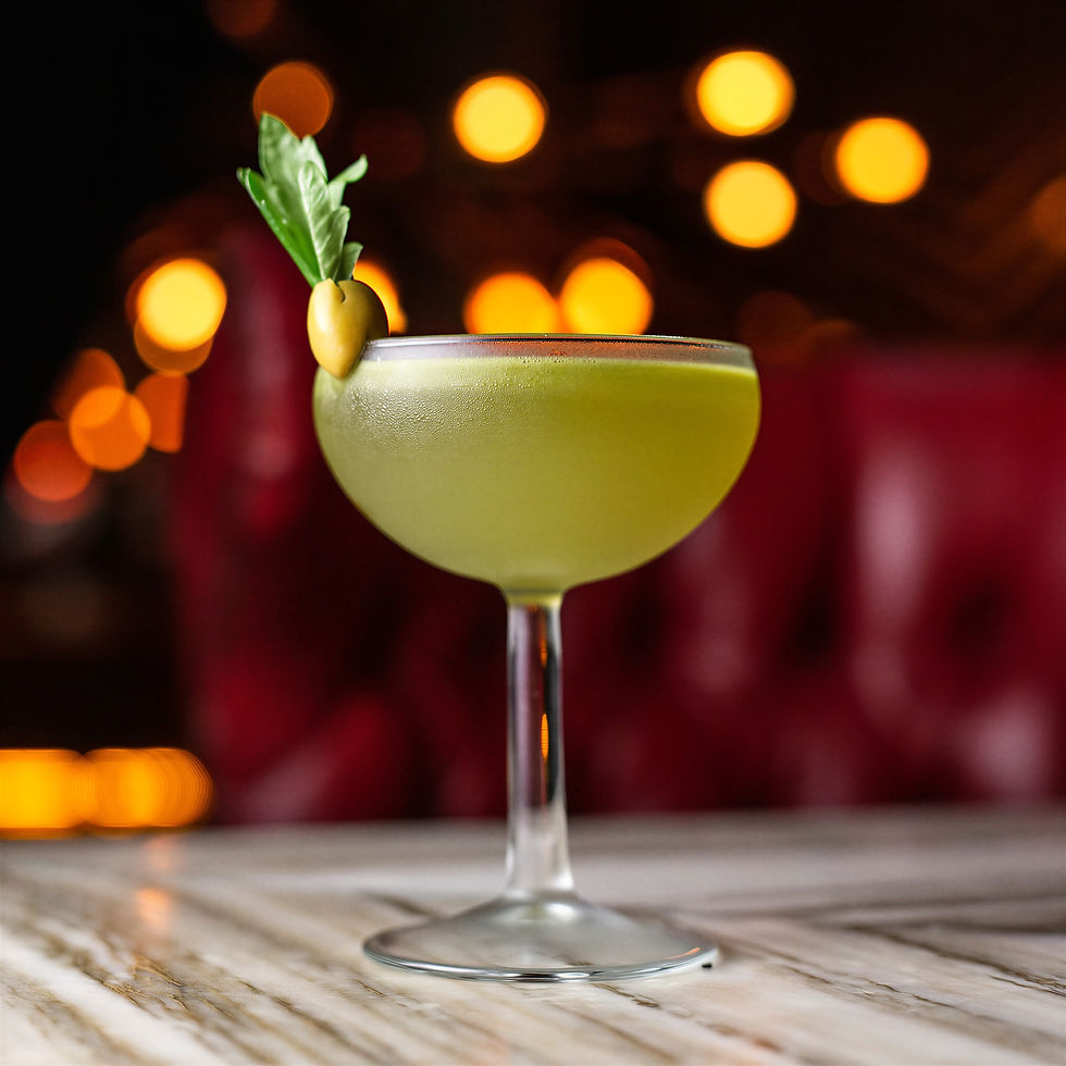 Green cocktail in a glass garnished with an olive and leaf sits on a marble table. Warm bokeh lights and red background create a cozy mood.