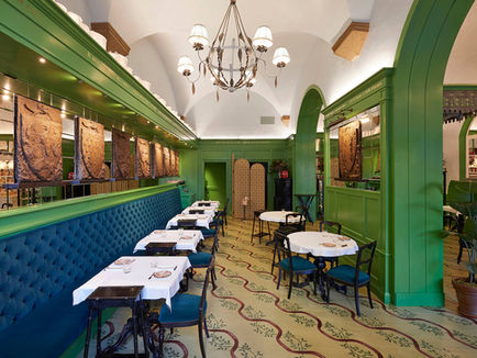 Elegant restaurant with green walls, ornate lighting, blue seating, and white tablecloths. Art and plants adorn the space, creating a cozy vibe.