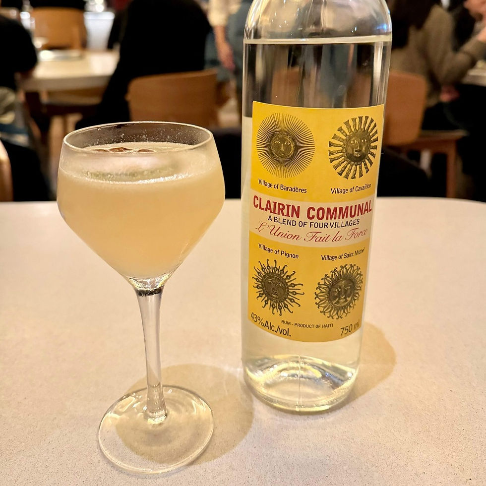 A glass of frothy cocktail alongside a bottle of 'Clairin Communal' rum, showcasing its label which mentions a blend from four villages in Haiti, set in a vibrant restaurant ambiance.