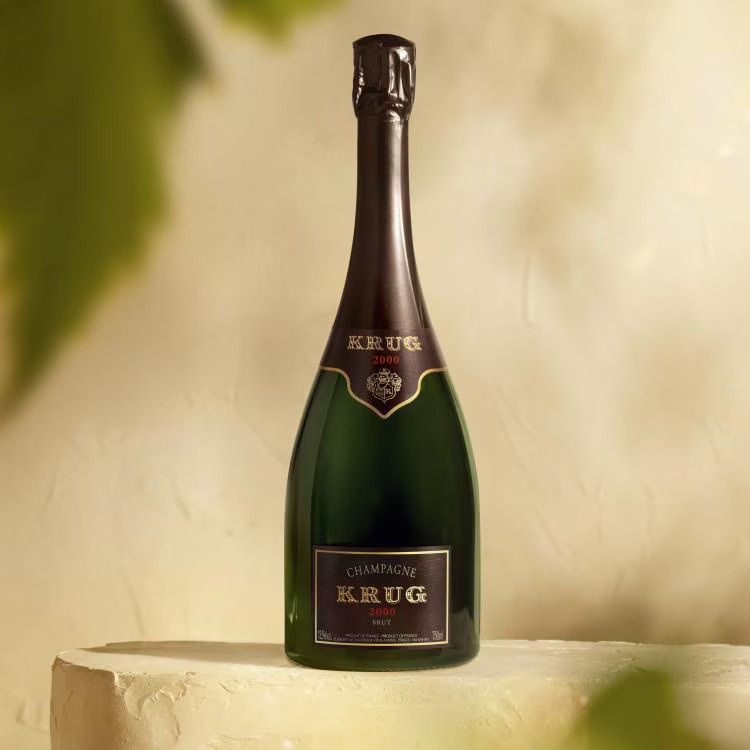 Champagne bottle labeled "Krug 2000" on a beige surface. Soft lighting and leafy shadows create an elegant, sophisticated mood.