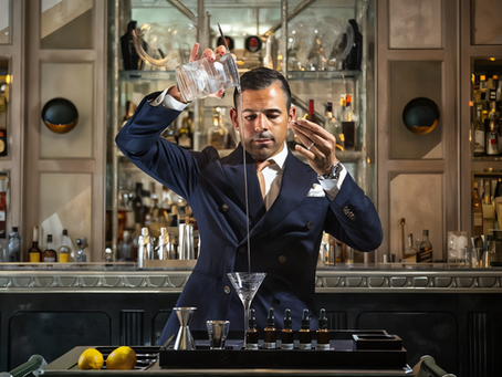 Bartender in a suit pours liquid from a high position into a martini glass in a stylish bar setting. Lemons and bottles are visible.