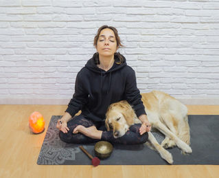 A woman in a black hoodie sits on a yoga mat in a light-colored room and meditates with her dog.