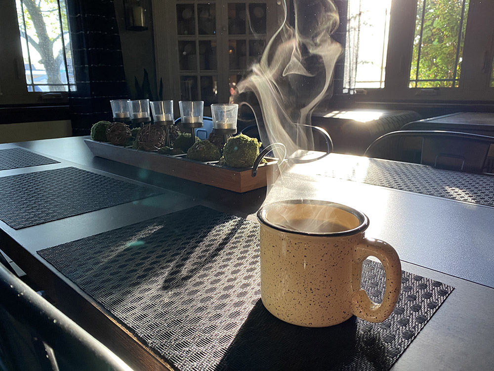 A cup of tea rests on a table and steam is visible in the sunlight coming through the window.
