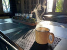 A cup of tea rests on a table and steam is visible in the sunlight coming through the window.