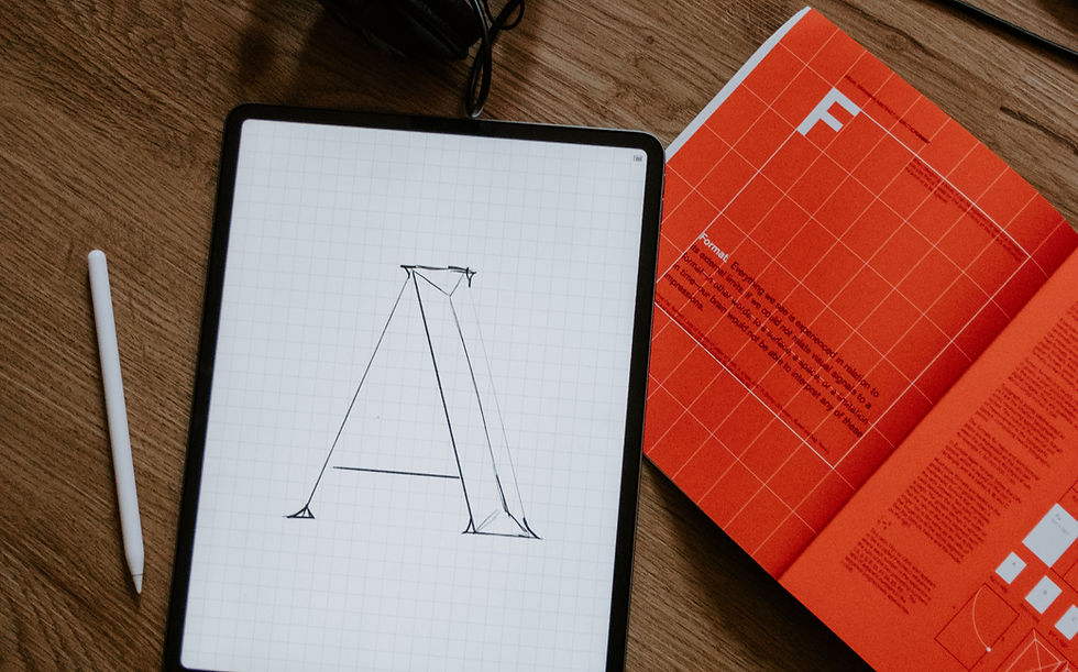 Tablet z rysunkiem litery "A" na tle w kratkę, obok biały rysik i czerwona książka z tekstem na drewnianym biurku.