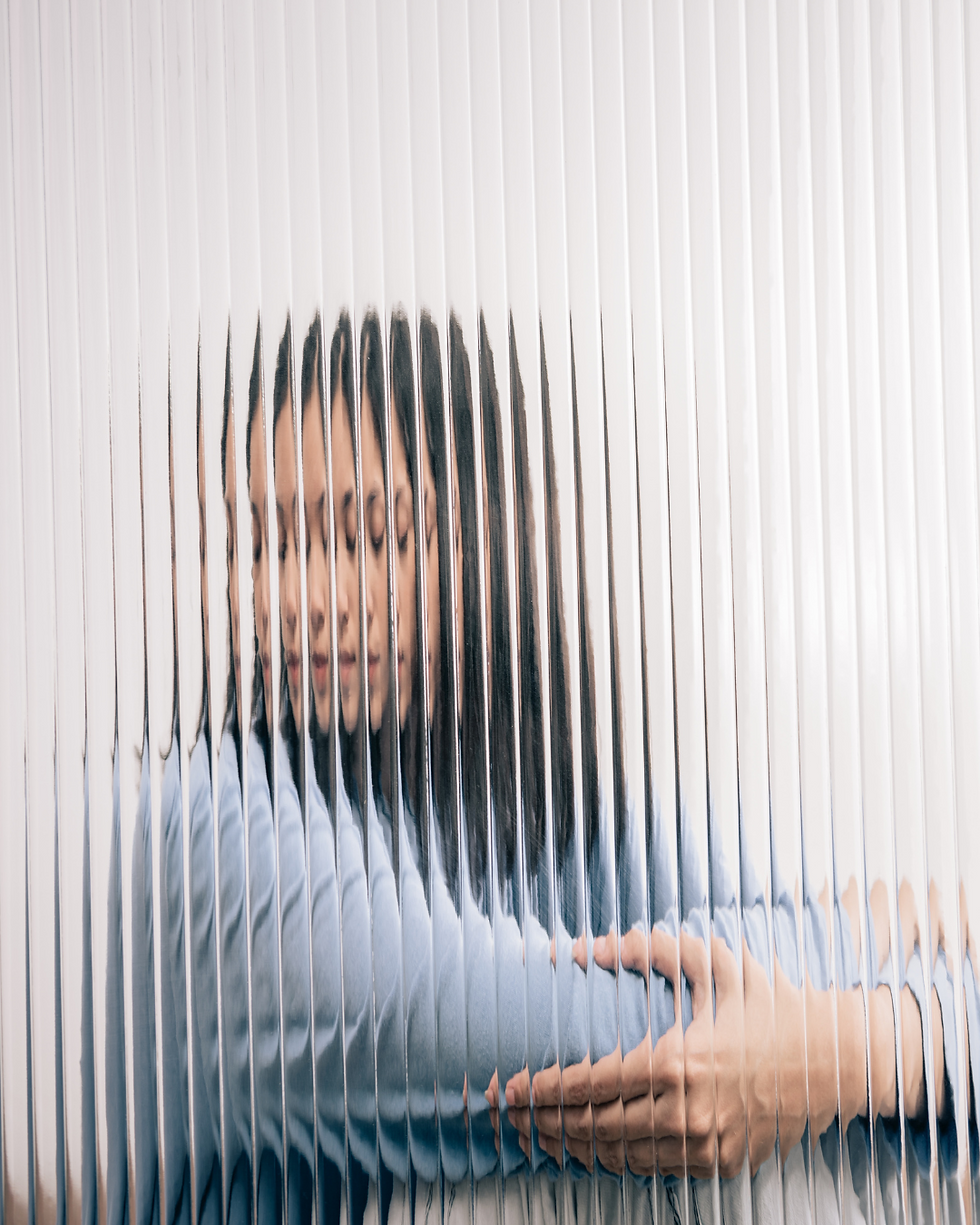 Femme en pull bleu vue à travers une paroi en verre ondulé, créant un effet de distorsion et de duplication. Elle ferme les yeux et semble s’enlacer, évoquant l’introspection, le rapport à soi et la perception déformée de l’image corporelle.