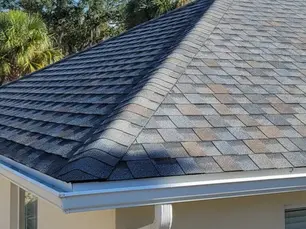A house with a shingle roof in shades of gray and brown, gutters visible, surrounded by green trees under a clear blue sky.