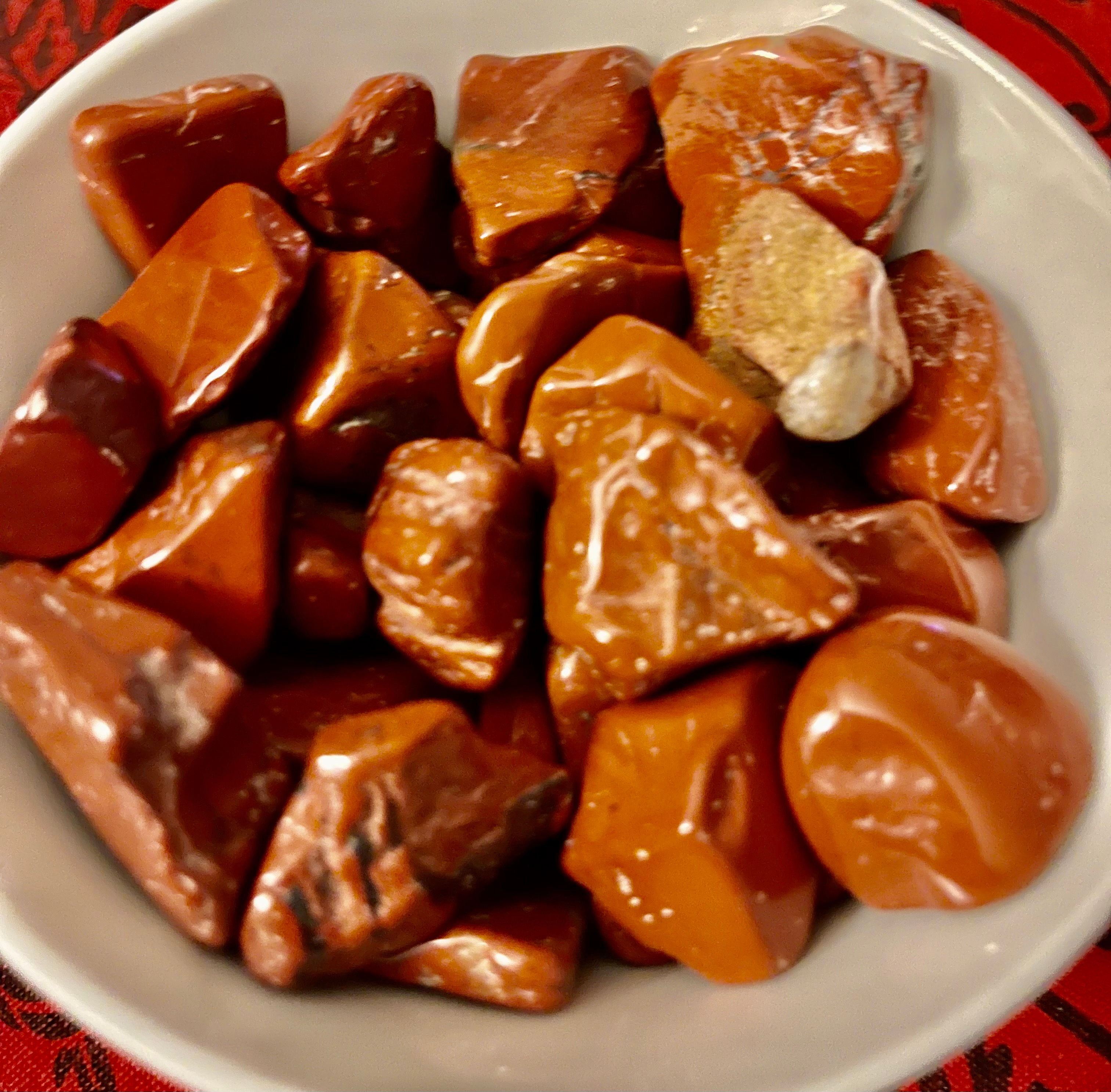 a large number of medium Red Jasper crystals, ranging greatly in color and size, sits in a white dish above a red table throw.