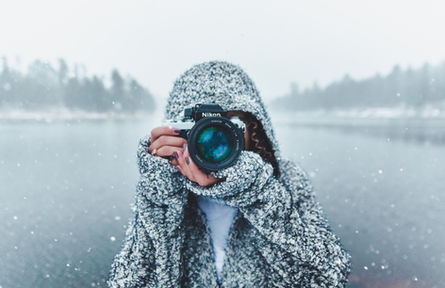 Hooded photographer aims a Nikon camera at the viewer beside a snowy, misty lake, creating a focused winter mood.