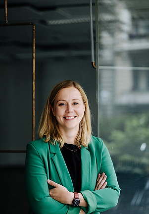 Portrait of Janine Langlotz in a green blazer with a dark shirt. She is standing in front of a shop window and smiles.