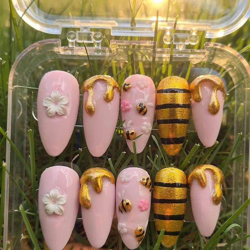 Luxury blush pink press-on nails with gold honey drip accents, bee details, and floral embellishments displayed in grass with warm golden sunlight.