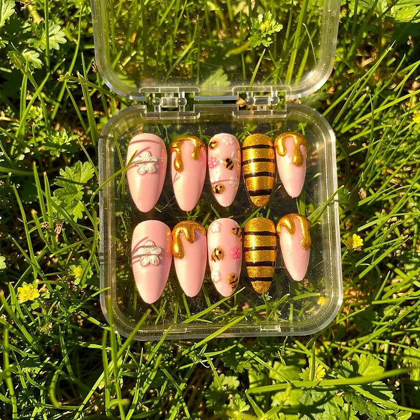 Luxury blush pink press-on nails with gold honey drip accents, bee details, and floral embellishments displayed in grass with warm golden sunlight.