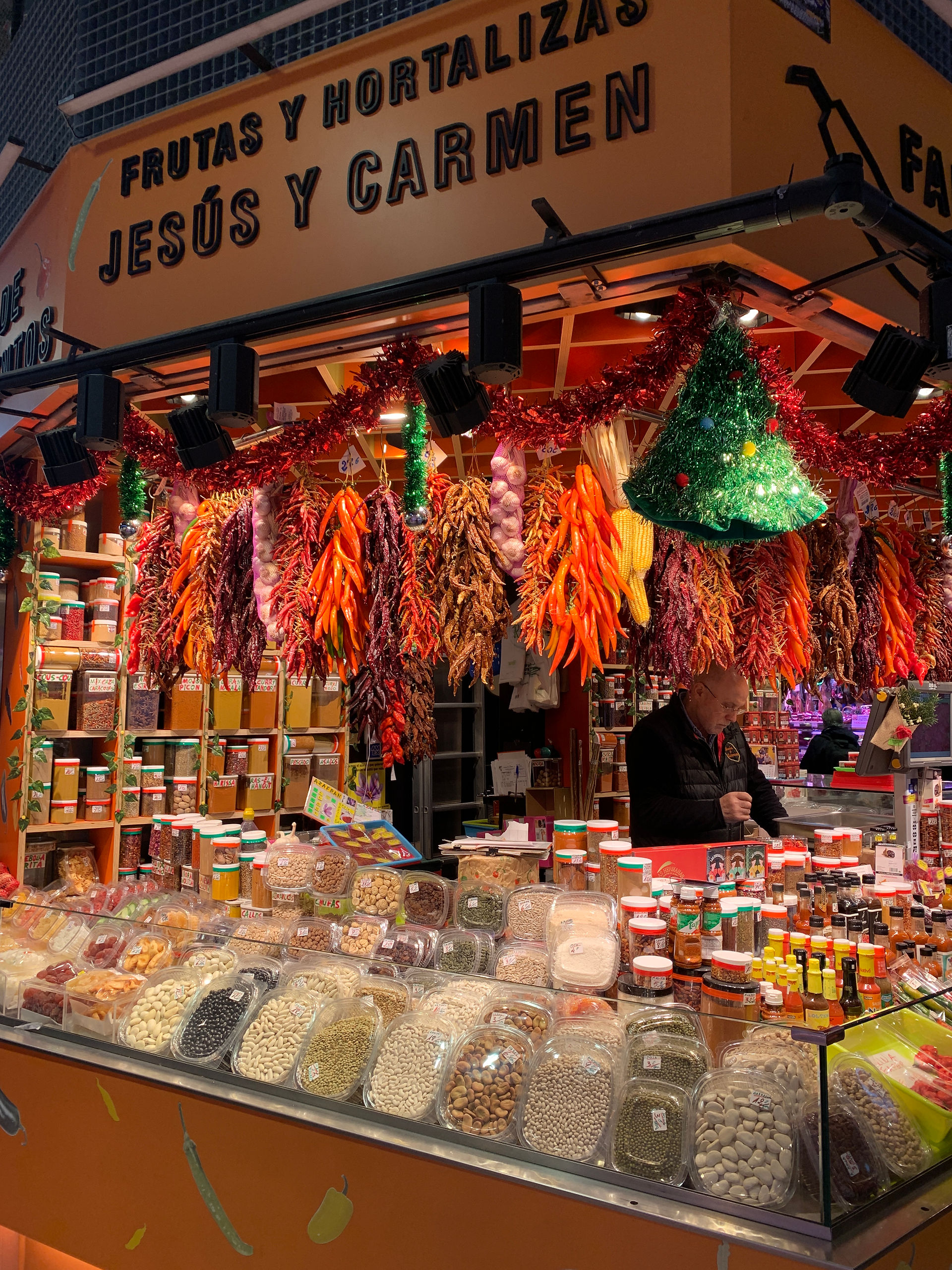 A market stall showcases a wide variety of beans, spices, and hanging dried peppers. The stall is festively decorated with garlands and a small green Christmas tree. A vendor in dark clothing stands behind the counter, surrounded by jars and packets of goods.