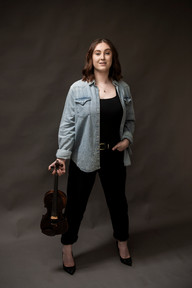 Standing portrait of Georgina Mabey in a denim jacket, holding her violin, looking directly at the camera.