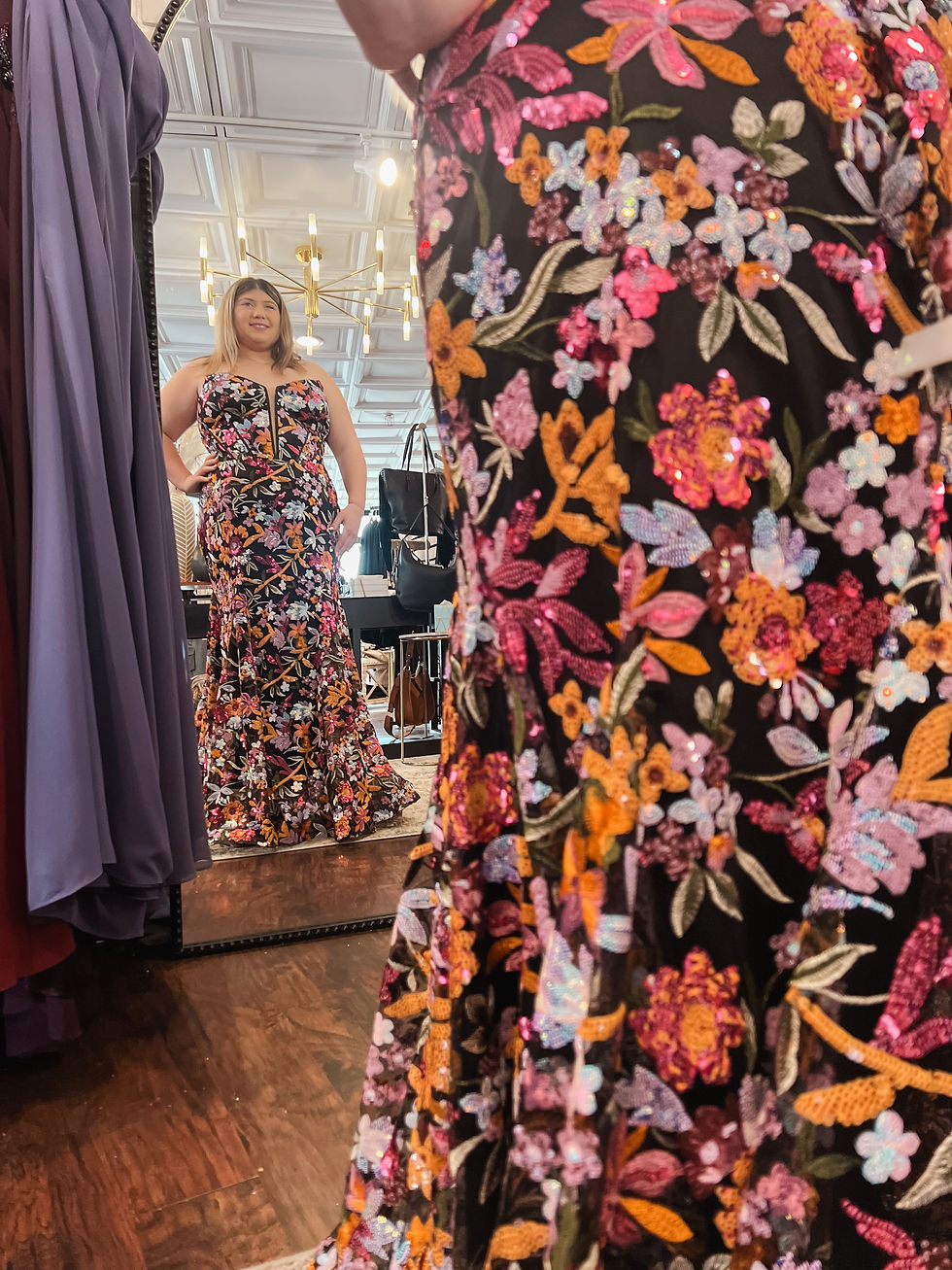 Woman in a floral dress admires her reflection in a store mirror. The dress is colorful and sequined. Background shows other dresses and lighting.